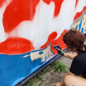 U27 Streetart Stipendium Landsberg am Lech 2025 zur Förderung der Kreativität von jungen Heranwachsenden und nachhaltigen Verschönerung des öffentlichen Raumes. Träger Kreisjugendring Landsberg. Konzept und Umsetzung Erwa.One. Jeanne streicht mit dem Pinsel den Hintergrund des Streetart-Motivs auf einen Container in Blau.