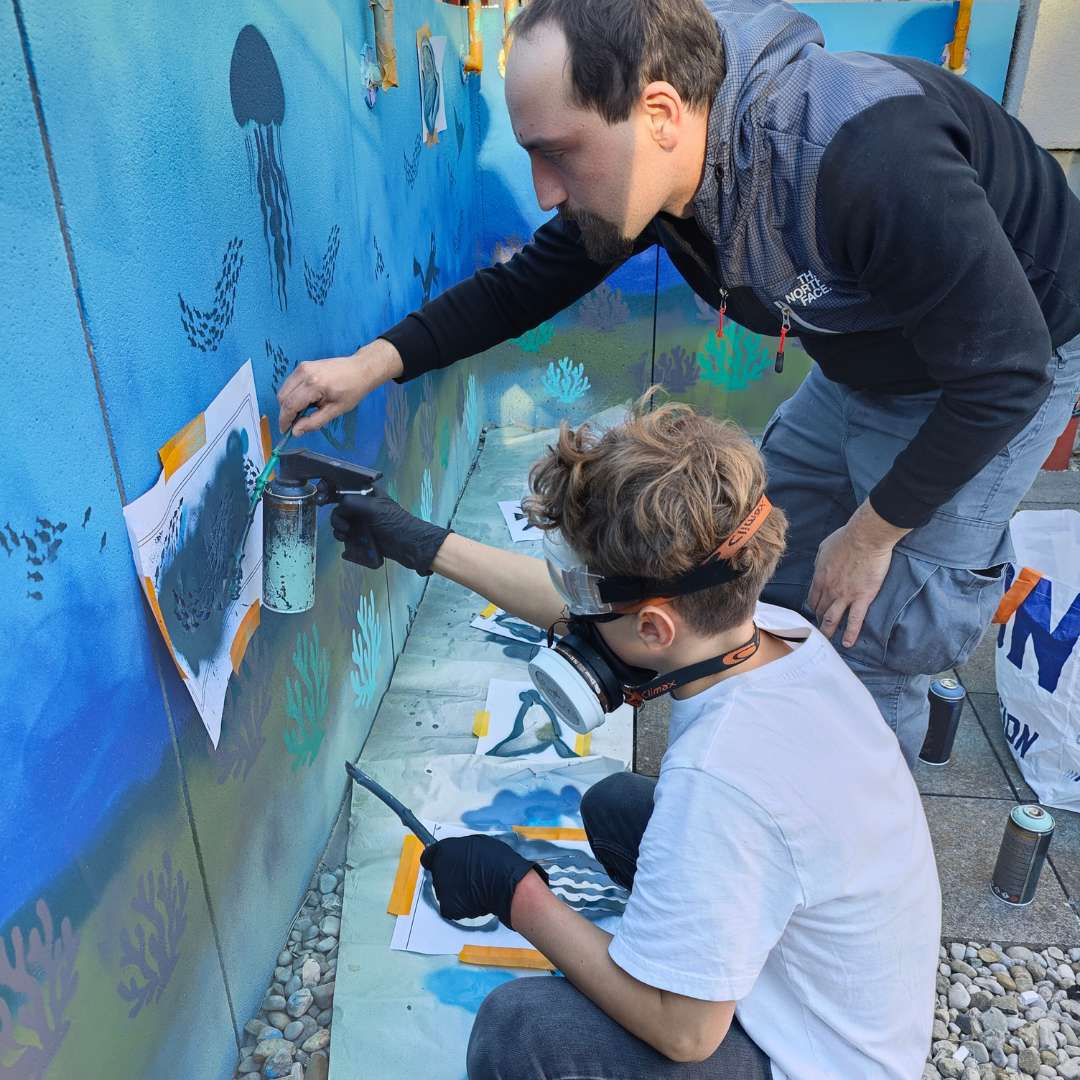 Kunstprojekt offene Jugendarbeit in Pöcking am Starnberger See. Thema des Streetart-Projektes "Unterwasserwelt" auf einer Betonmauer am Schwimmbad von Pöcking. Künstler Erwa.One unterstützt einen Teilnehmer beim sprühen einer Schablone.