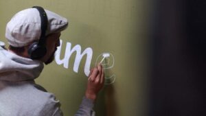 B2B Auftragsgestaltung für einen Gastronomiebetrieb Fiume in Landsberg am Lech. Künstler Erwa.One füllt mit dem Pinsel den Buchstaben "e" auf der Wand mit weißer Farbe aus. Er trägt Kopfhörer und eine graue mütze.