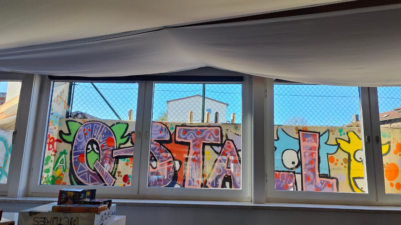 Kunstprojekt offene Jugendarbeit in Pöcking am Starnberger See. Thema des Streetart-Projektes "Unterwasserwelt" auf einer Betonmauer am Schwimmbad von Pöcking. Einführungssprühkurs im Jugendtreff. Blick durchs Fenster auf Wand mit Graffiti Schriftzug Q-Stall.