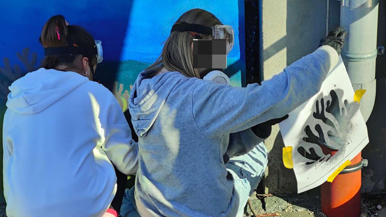 Kunstprojekt offene Jugendarbeit in Pöcking am Starnberger See. Thema des Streetart-Projektes "Unterwasserwelt" auf einer Betonmauer am Schwimmbad von Pöcking. Zwei Mädchen sprühen auf eine Wand. Die linke Akteurin legt eine Korallen-Schablone zur Seite. Beide tragen eine Schutzmaske und Schutzbrillen
